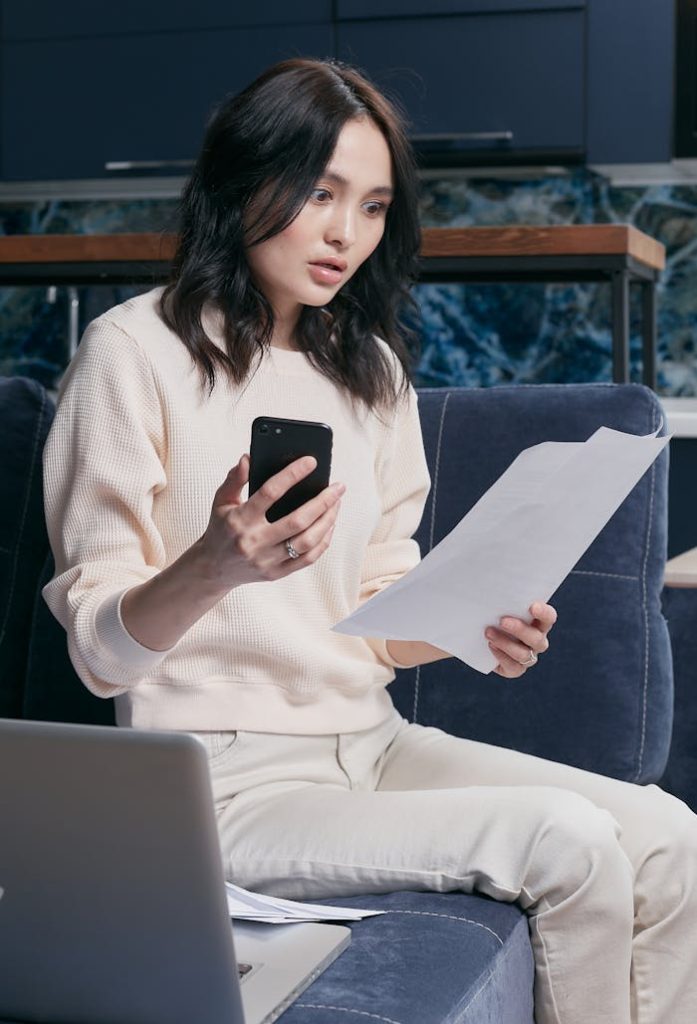 The Art of Drawing Readers In: Your attractive post title goes here A young woman uses her smartphone and laptop to manage finances, appearing focused and concerned.